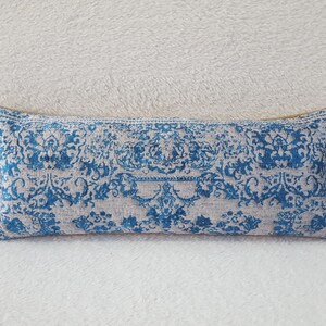 May include: A rectangular decorative pillow with a blue and beige floral pattern. The pillow has a soft, textured appearance and is placed on a white, fluffy surface. The design features intricate, stylized floral motifs.