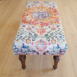 Puede incluir: Un otomano con estampado colorido y patas de madera. El otomano tiene un dise&ntilde;o floral con tonos de azul, rosa, amarillo y naranja.