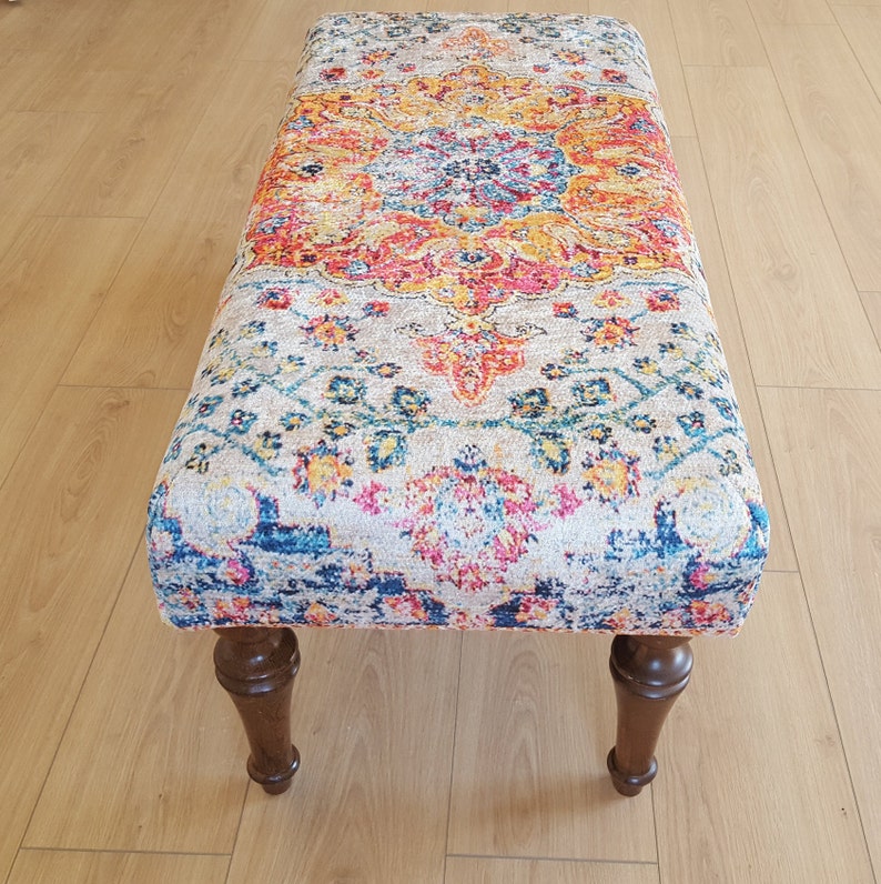 Puede incluir: Un otomano con estampado colorido y patas de madera. El otomano tiene un dise&ntilde;o floral con tonos de azul, rosa, amarillo y naranja.