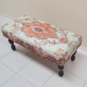 May include: A light green and orange patterned ottoman with dark brown wooden legs. The ottoman is sitting on a white tiled floor.