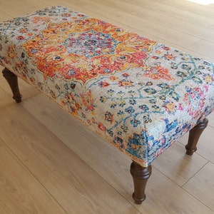 Puede incluir: Un otomano con estampado colorido sobre un fondo beige y patas de madera. El estampado presenta tonos de rojo, naranja, amarillo, azul y verde.