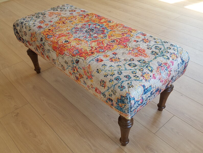 Puede incluir: Un otomano con estampado colorido sobre un fondo beige y patas de madera. El estampado presenta tonos de rojo, naranja, amarillo, azul y verde.