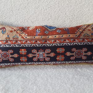 May include: A long rectangular pillow with a colorful patterned design. The pillow features a red, blue, and white geometric pattern reminiscent of a traditional rug.
