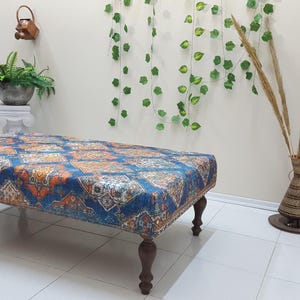Peut inclure: Un ottoman bleu et orange &agrave; motifs avec quatre pieds en bois. L'ottoman est pos&eacute; sur un sol en carrelage blanc.