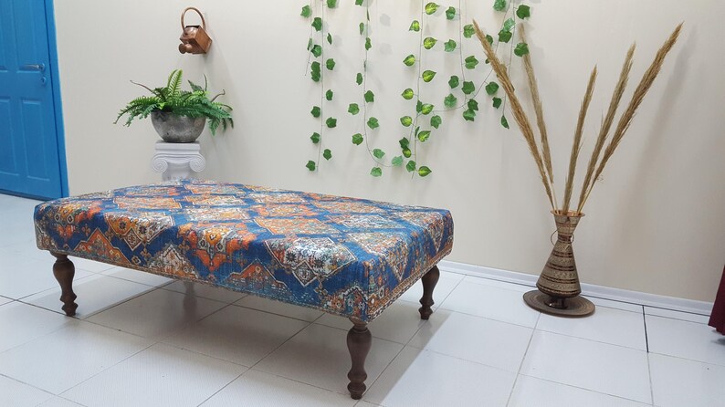Peut inclure: Un ottoman bleu et orange &agrave; motifs avec quatre pieds en bois. L'ottoman est pos&eacute; sur un sol en carrelage blanc.