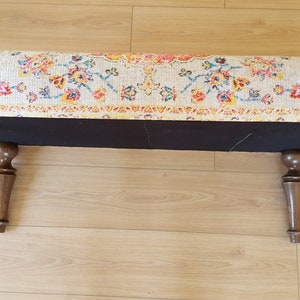 Puede incluir: Un otomano rectangular con una parte superior de tela con estampado floral colorido y cuatro patas de madera marr&oacute;n oscuro. El otomano est&aacute; sobre un suelo de madera clara.