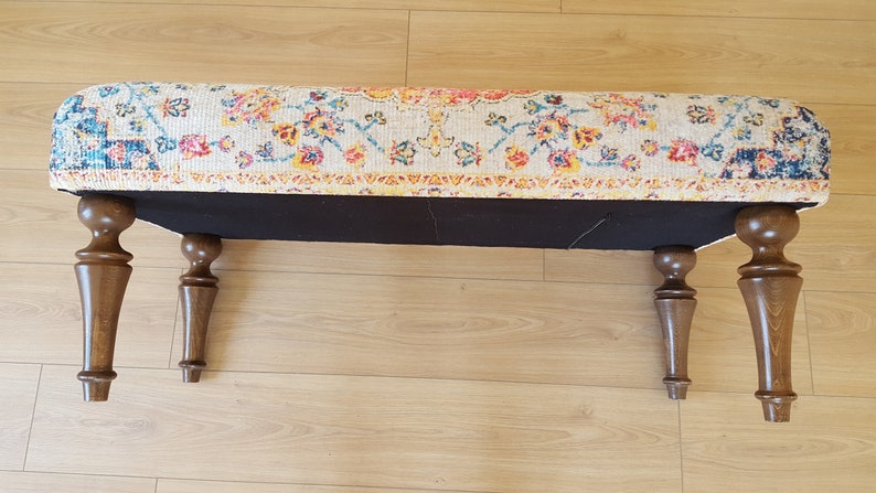 Puede incluir: Un otomano rectangular con una parte superior de tela con estampado floral colorido y cuatro patas de madera marr&oacute;n oscuro. El otomano est&aacute; sobre un suelo de madera clara.
