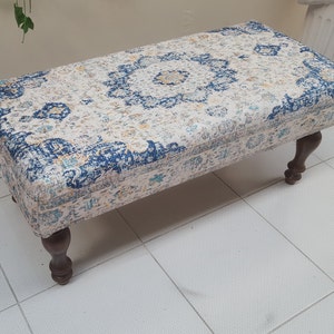 May include: A white ottoman with a blue and gold patterned fabric top and four dark wood legs.