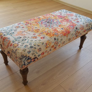 Puede incluir: Un otomano rectangular con una parte superior de tela estampada de colores y cuatro patas de madera. La tela presenta un gran dise&ntilde;o floral intrincado en tonos de rojo, naranja, amarillo, azul y verde.