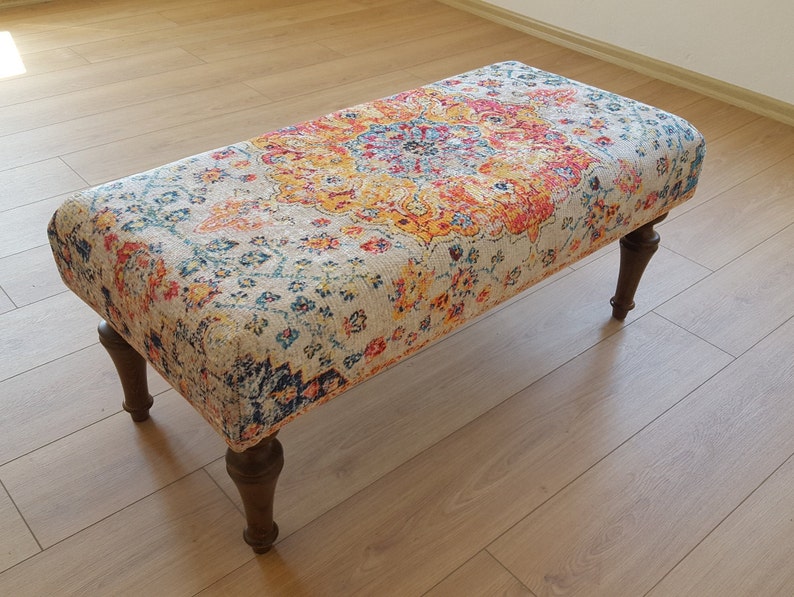 Puede incluir: Un otomano rectangular con una parte superior de tela estampada de colores y cuatro patas de madera. La tela presenta un gran dise&ntilde;o floral intrincado en tonos de rojo, naranja, amarillo, azul y verde.