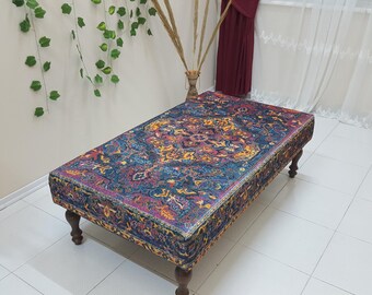 Puede incluir: Un otomano colorido y estampado con un fondo azul oscuro y diseños florales intrincados en tonos rojos, amarillos y dorados. El otomano tiene un marco de madera con cuatro patas.
