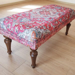 May include: A red, blue and white patterned ottoman with wooden legs. The ottoman is sitting on a light wood floor.
