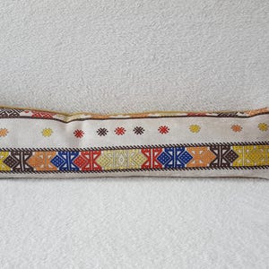May include: A long, rectangular pillow with a white background and colorful geometric patterns in shades of red, yellow, blue, and brown. The pillow is made of a soft, woven fabric.