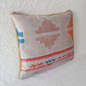 May include: A rectangular cushion with a beige background and a colourful geometric pattern in shades of orange, red, blue, and white. The cushion has a yellow border.