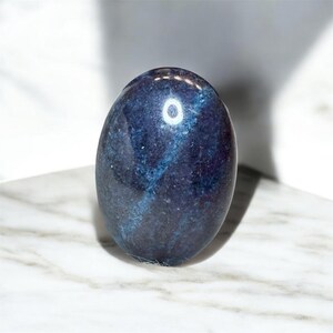 May include: A polished, oval-shaped stone with a deep blue hue and lighter blue veining. The stone is smooth and reflective, resting on a white and grey marble surface. The overall aesthetic is one of natural beauty and elegance.