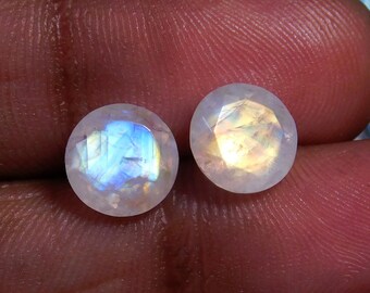 May include: Two round, iridescent moonstone cabochons with a white, blue, and gold color. The stones are faceted and have a smooth, polished finish.