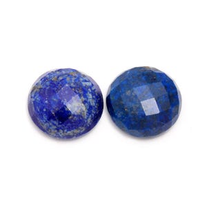 May include: Two round, faceted lapis lazuli gemstones. The gemstones are a deep blue color with flecks of gold and white. The facets catch the light, creating a sparkling effect. The gemstones are set against a white background.