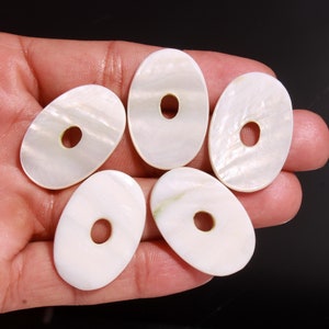 Può includere: Cinque perline ovali di madreperla bianca con un foro al centro. Le perline sono lisce e hanno una leggera lucentezza iridescente.