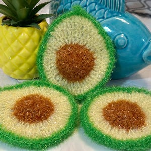 May include: Three handmade crochet scrubbers shaped like avocado halves. Each scrubber features a bright green outer edge, a pale yellow-green center, and a textured, metallic-toned brown circular pit in the middle, crafted from scrubby yarn.