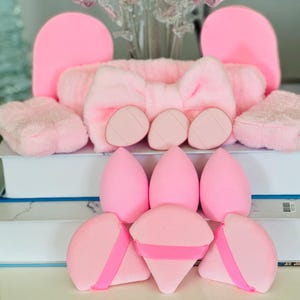 May include: A collection of pink beauty accessories, including a headband with a bow, makeup sponges, and face cleansing pads. The items are arranged on top of white books, creating a clean and organized display. The soft, plush textures and pastel color palette give a feminine aesthetic.