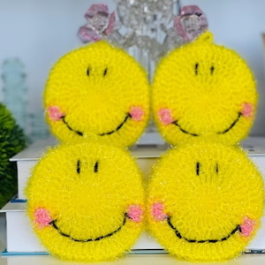 May include: Four yellow smiley face scrubbies with pink cheeks and black smiles. These round, textured scrubbers are stacked on a white surface. The scrubbies are a cheerful addition to any kitchen or bathroom.