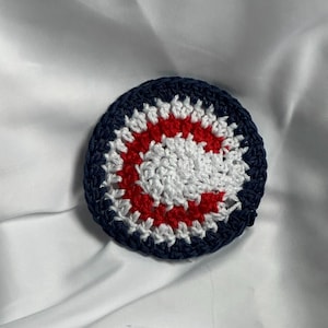 May include: A round, crocheted coaster with a navy blue border, a red center ring, and a white inner circle. The coaster is handmade and sits on a white, silky surface. The design is reminiscent of a target.