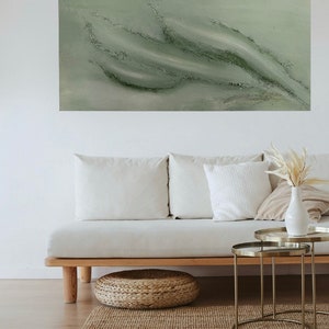 Könnte beinhalten: Ein grünes abstraktes Gemälde mit weißen Akzenten hängt über einem weißen Sofa mit Kissen. Das Sofa steht vor einem hellen Holzboden und einem gewebten Teppich. Zwei goldene Beistelltische aus Metall stehen neben dem Sofa.