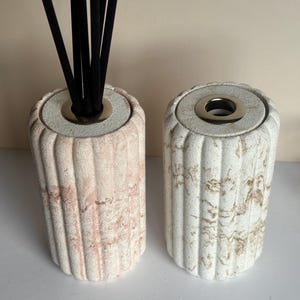 May include: Two decorative reed diffuser holders. One is pink and the other is white with brown marbling. Both have a cylindrical shape with vertical grooves and a silver-coloured ring at the top. Black reeds are inserted into the pink holder.