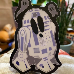 May include: A white and blue robot with black eyes and a ghost-like outline. Two small ghosts are on either side of the robot.