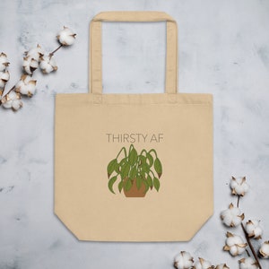 Può includere: Una borsa tote beige con un'illustrazione di una pianta appassita e il testo "THIRSTY AF" stampato su di essa.
