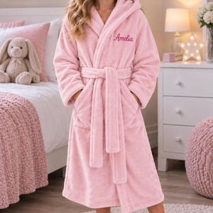 May include: A soft, light pink plush bathrobe featuring a hood, long sleeves, and a matching tie belt at the waist. The robe has two front pockets and the name Amelia embroidered in dark pink cursive script on the upper left chest area.