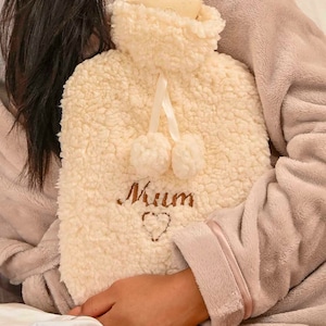 Puede incluir: Una botella de agua caliente blanca y esponjosa con una cinta de color crema y dos pompones. La botella tiene la palabra "Mamá" bordada con un pequeño corazón.