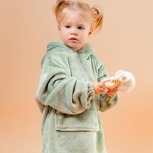May include: A young child wearing a soft, sage green hooded blanket with a front pocket. The child is holding a white and tan toy. The background is a solid peach colour.