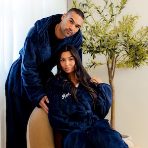 Pode incluir: Duas pessoas usando roupões com capuz azul marinho combinando. O roupão da pessoa em primeiro plano tem a palavra "Hot" bordada em branco. Os roupões parecem ser feitos de um material macio e felpudo.