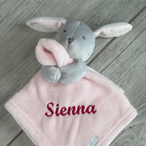 May include: Pink baby blanket with a gray bunny attached. The blanket is embroidered with the name "Sienna".