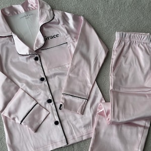May include: Pink satin pajama set with black trim and buttons. The top has a breast pocket and the name "Grace" embroidered on it. The label reads "Lulabay Kids 5-6Y". The set includes a long-sleeved button-down shirt and matching pants.
