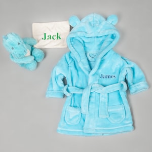 Può includere: Una vestaglia con cappuccio di colore azzurro chiaro con cintura e tasche. La vestaglia è ricamata con il nome "James". Sullo sfondo c'è un asciugamano bianco con il nome "Jack".