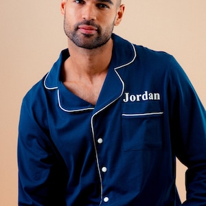 May include: Navy blue pajama top with white piping and buttons. The word "Jordan" is embroidered on the chest pocket. The long-sleeved top is part of a sleepwear set.