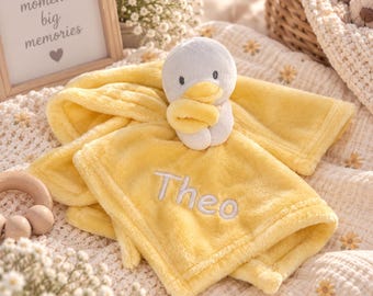 May include: A soft yellow plush security blanket featuring an attached stuffed duck head and wings. The fabric is embroidered with the name Theo in white thread. The blanket has a fuzzy texture and is designed for comfort and tactile engagement.