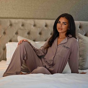 May include: Mauve pyjamas, comprising a long-sleeved top and trousers. The top features a breast pocket and the name "Eleanor" embroidered in black. The soft, flowing fabric of the pyjamas is displayed on a bed.