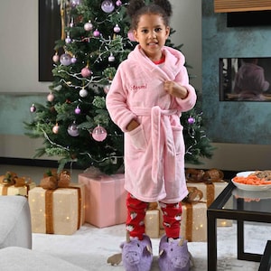 May include: A young person wearing a pink hooded robe with the name "Stormi" embroidered on the chest, red patterned pajamas, and purple dragon slippers. A Christmas tree with ornaments and wrapped gifts are in the background.