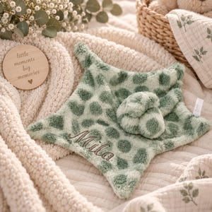 Peut inclure: Une couverture de sécurité en peluche en forme d'étoile, vert sauge doux avec un motif à pois vert foncé. Le tissu est duveteux et texturé. Le prénom Mila est brodé en écriture cursive marron foncé sur le devant. Une petite boule est fixée.
