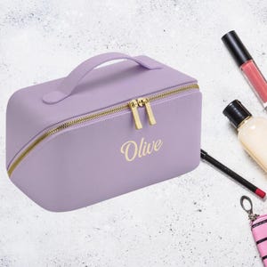 May include: A lavender-coloured zippered makeup bag with a gold zipper and handle. The bag has the name "Olive" written in gold lettering on the front.