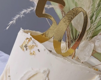 May include: A white cake with gold leaf and a gold '50' topper. The cake is decorated with dried pampas grass and other dried flowers.