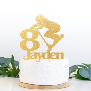 May include: Gold glitter cake topper featuring a silhouette of a person riding a scooter, the number 8, and the name Jayden.