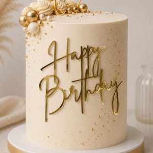 May include: A cylindrical cake with smooth cream-colored frosting, decorated with gold and white spheres, piped cream swirls, and small gold sprinkles. A gold acrylic cake topper reads Happy Birthday in cursive script, set on a white cake board.