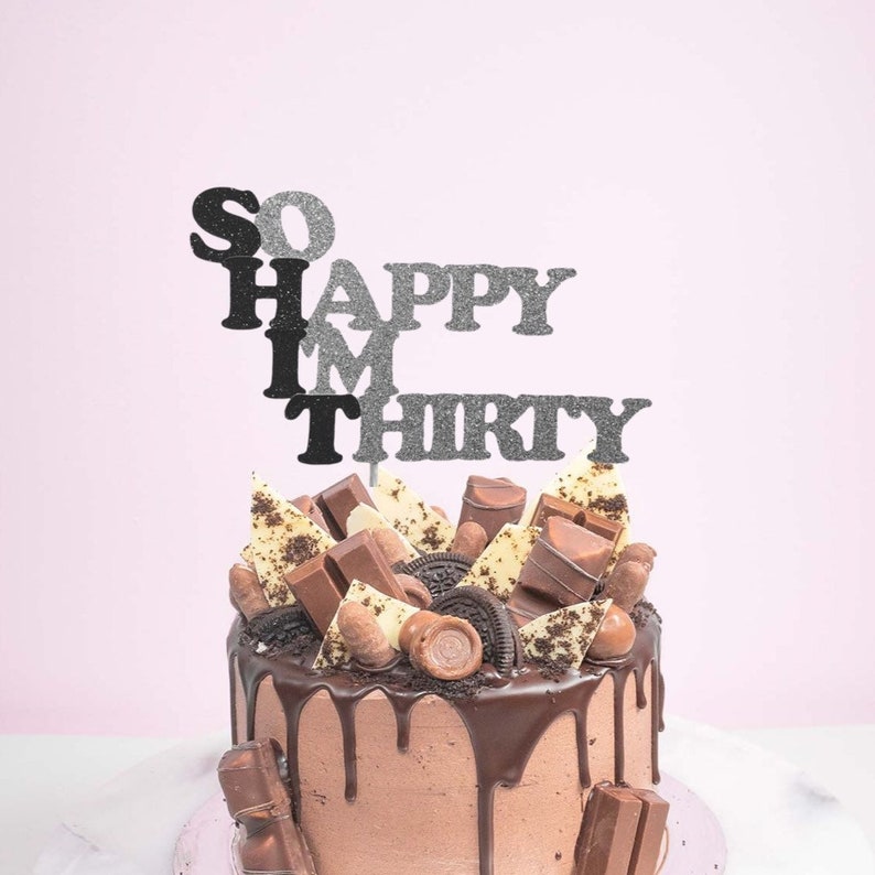 May include: A chocolate cake with a chocolate ganache drip and topped with chocolate candies, cookies, and a cake topper that reads "So Happy I'm Thirty".