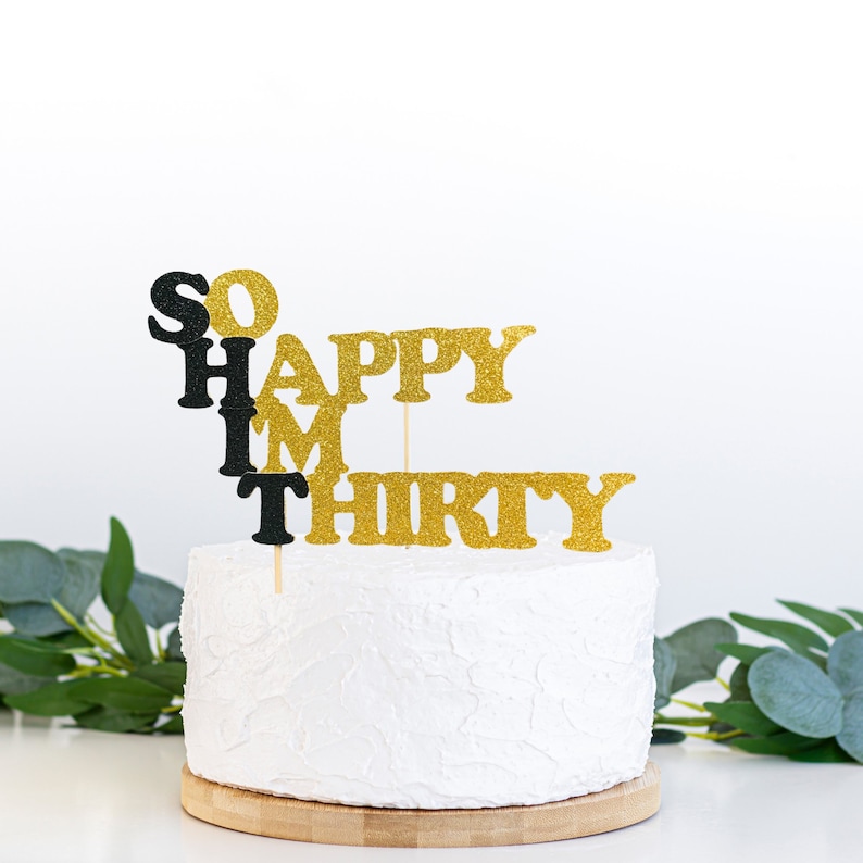 May include: White cake with gold glitter 'SO HAPPY I'M THIRTY' cake topper. The cake is on a wooden cake stand.