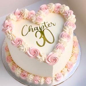 May include: A white heart-shaped cake decorated with pink frosting roses and gold lettering that says "Chapter 30".