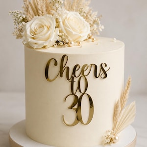 May include: A cylindrical cake with smooth cream-colored frosting, topped with white roses, dried grasses, and baby's breath. A gold-colored acrylic cake topper reads "Cheers to 30" in a script font. Small white pearls are scattered on the cake surface.